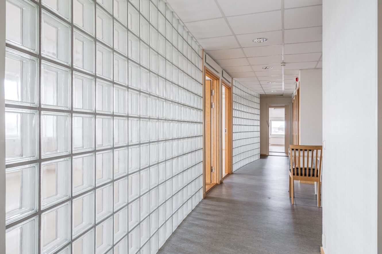 En korridor med en vägg av glasblock på ena sidan, trädörrar på den andra, en stol mot väggen och ett fönster i slutet av korridoren.