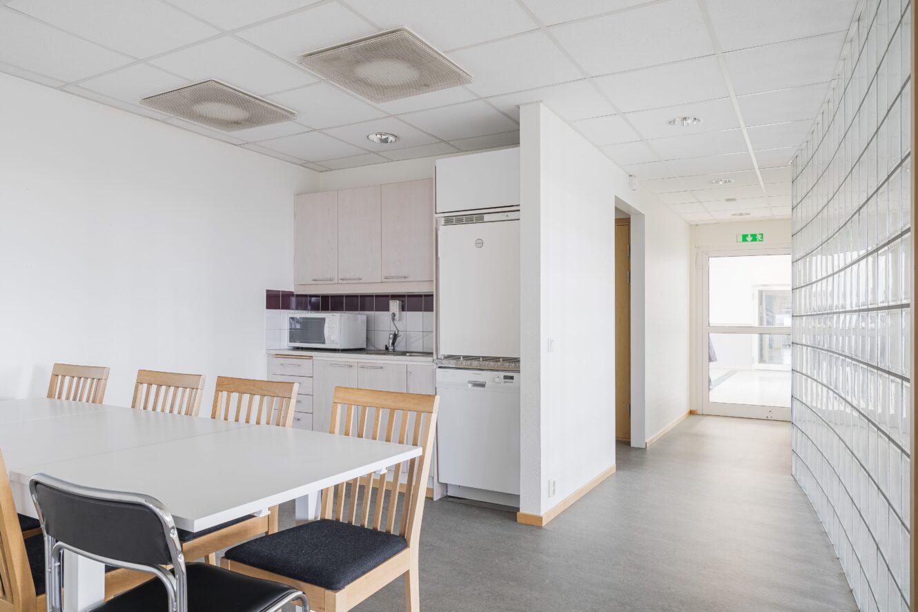 Ett litet kök med vita skåp, en mikrovågsugn och ett matbord med trästolar visas bredvid en hall med glasväggar och en synlig utgångsskylt.