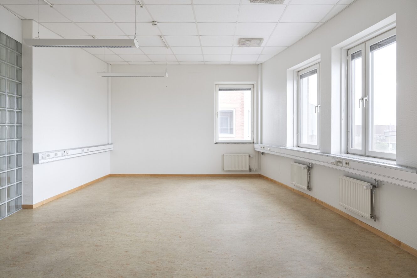 Tomt, omöblerat rum med vita väggar, stora fönster, ljust golv och takpaneler, med radiatorer och eluttag längs väggarna.