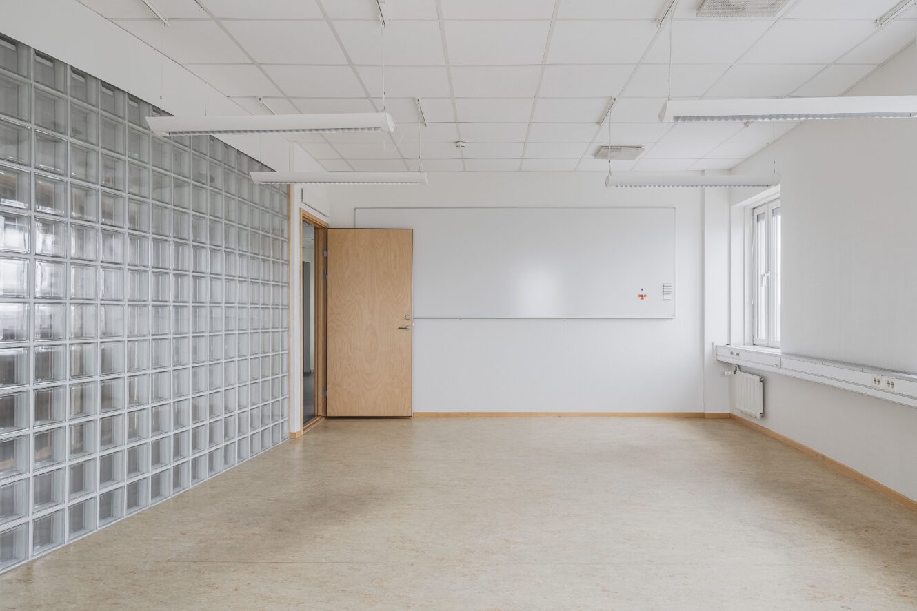 Tomt klassrum med en glasvägg, beige golv, whiteboard, takbelysning, en öppen trädörr och fönster till höger som släpper in naturligt ljus.