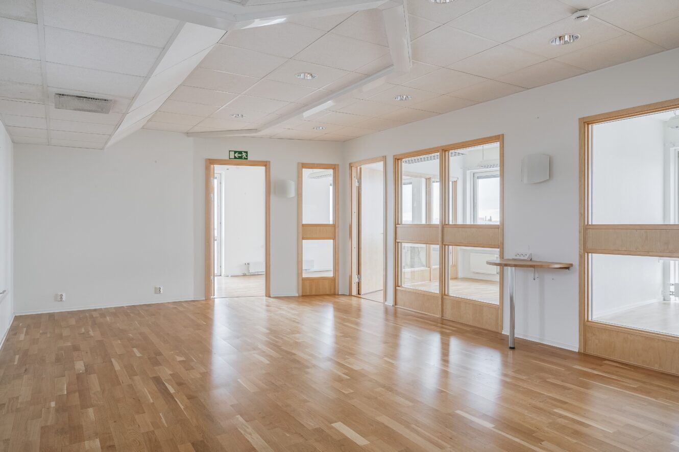 Rymligt tomt kontor med ljust trägolv, vita väggar, flera rum med glasväggar och taklampor.