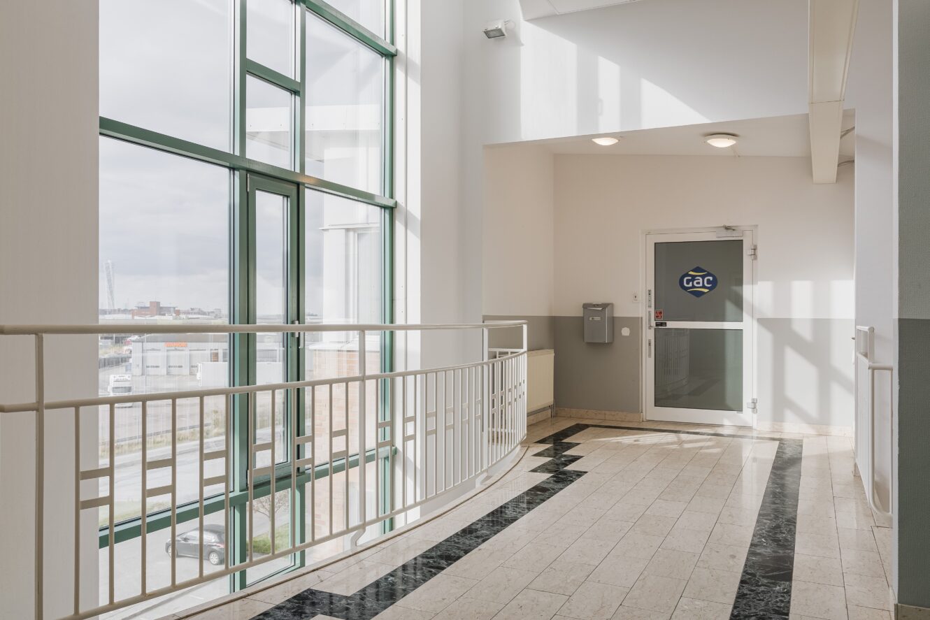 En korridor med stora fönster, ett vitt räcke, klinkergolv och en dörr märkt GAC i slutet. Utrymmet fylls av naturligt ljus.