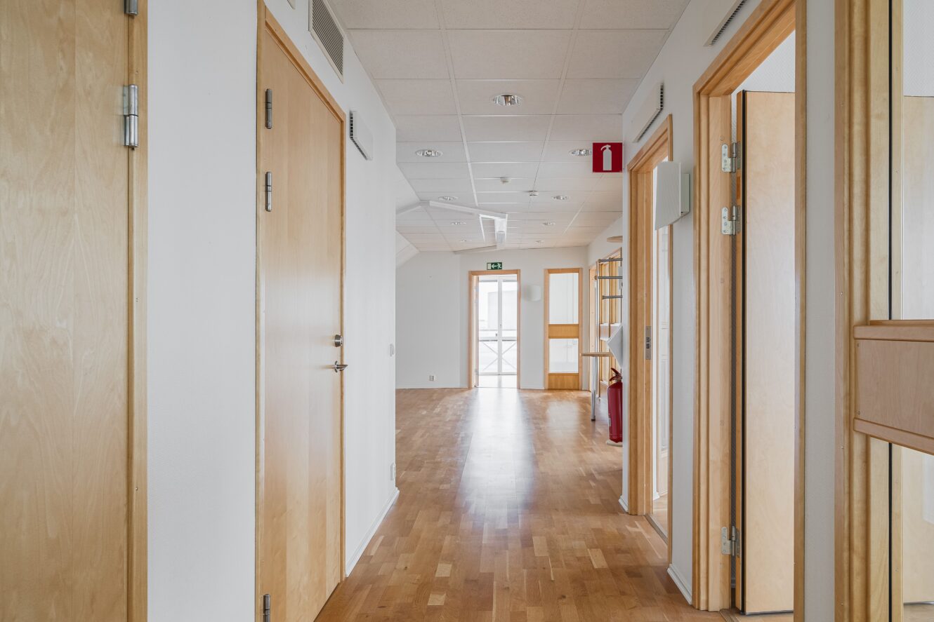 En hall med ljusa trädörrar, vita väggar och trägolv, som leder till en glasdörr i slutet med en nödutgångsskylt ovanför.