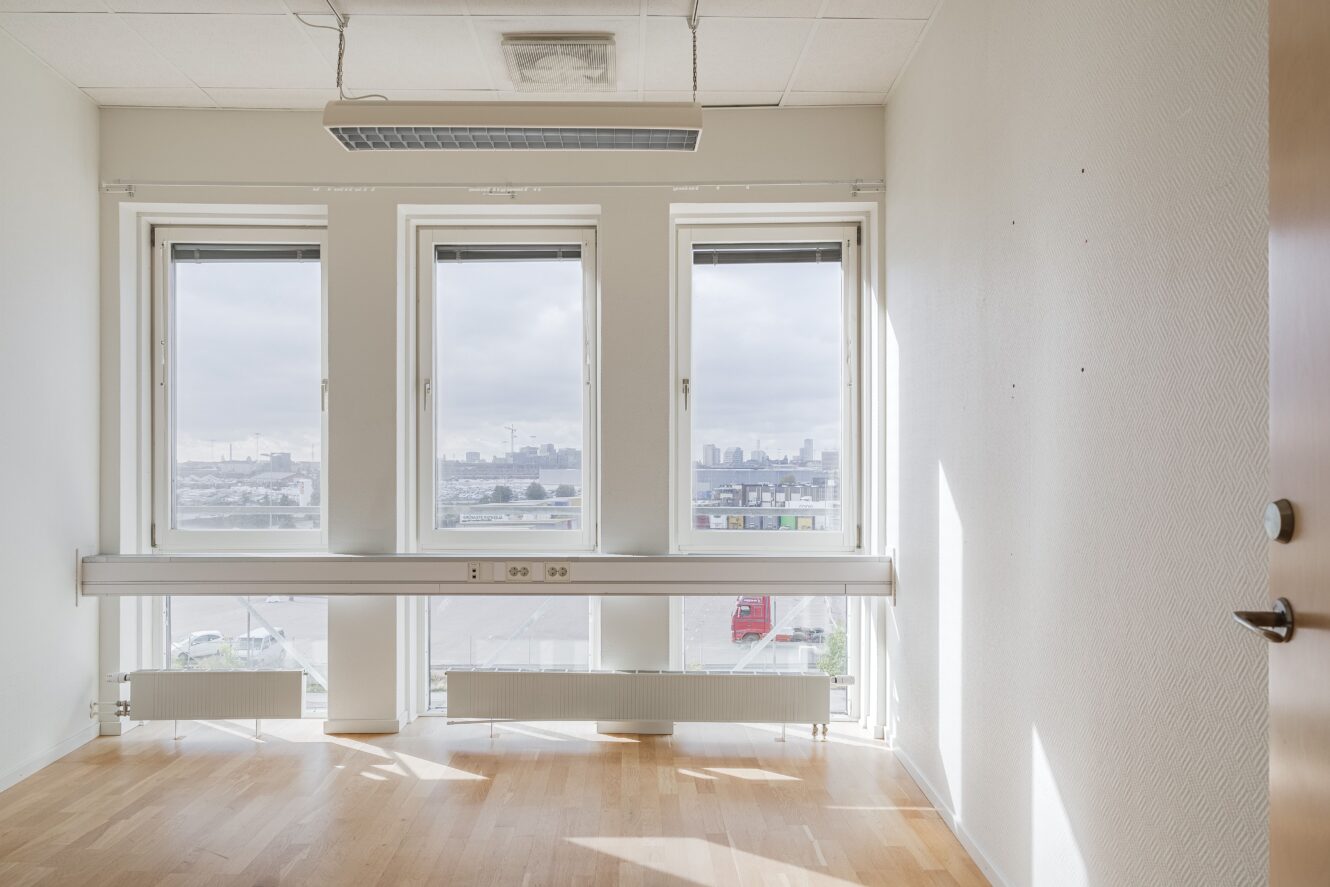 Tomt kontorsrum med tre stora fönster, vita väggar, ljust trägolv, takarmatur och utsikt över en stadssiluett i bakgrunden.