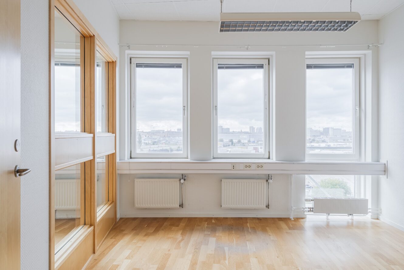 Tomt rum med ljust trägolv, tre stora fönster, vita väggar och två radiatorer under fönstren. En glasvägg med träinramning finns på vänster sida.