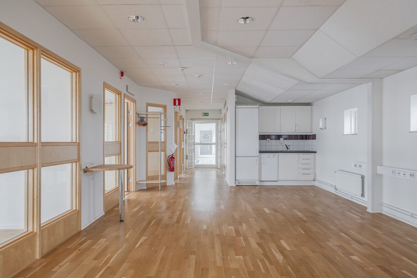 En ljus, tom hall med trägolv, stora fönster och ett pentryområde; dörrar och armaturer kantar väggarna.