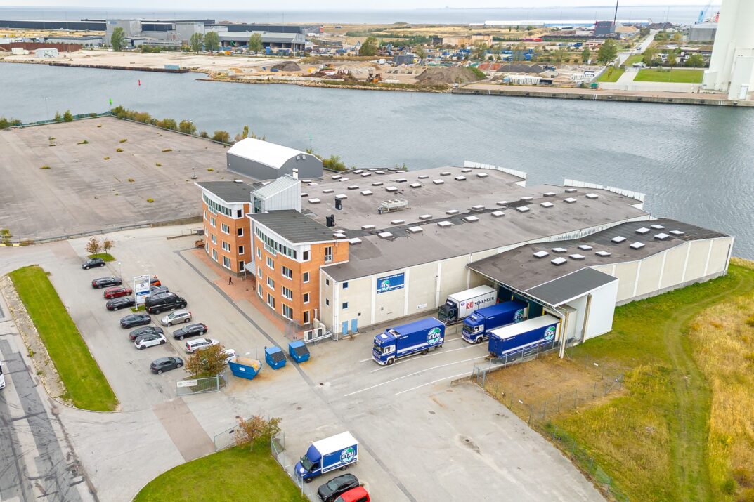 Flygfoto över en lagerlokal vid vattnet med flera parkerade bilar, budbilar och intilliggande tomma tomter.