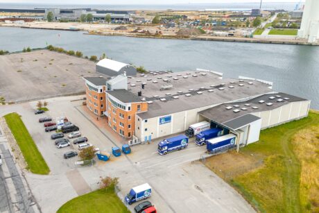 Flygfoto över en lagerlokal vid vattnet med flera parkerade bilar, budbilar och intilliggande tomma tomter.