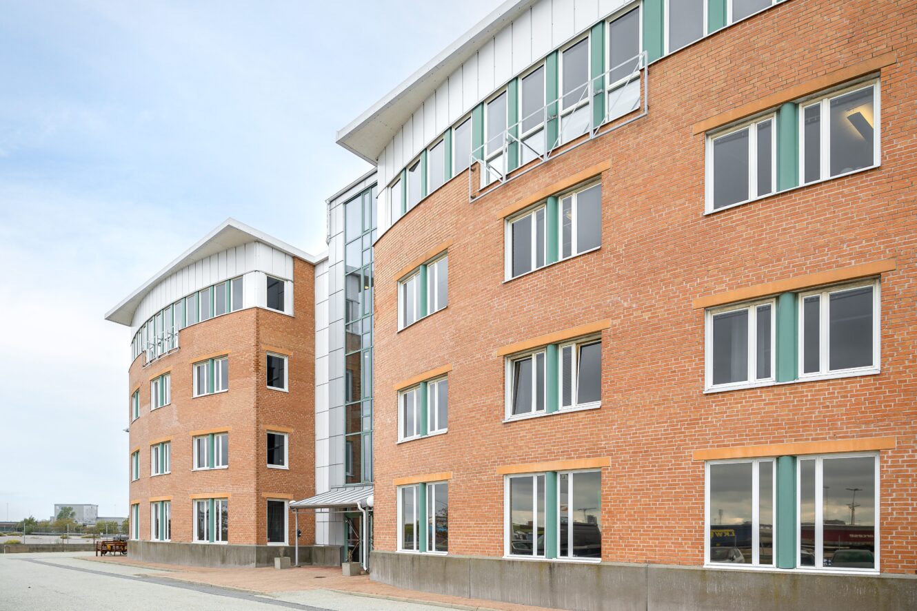 En modern kontorsbyggnad i tegel med flera våningar, stora fönster och en ingång i glas, fotograferad en klar dag.