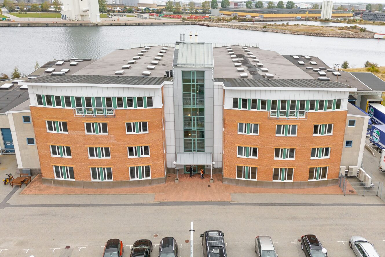 En fyra våningar hög kontorsbyggnad i tegel och glas vetter mot en parkeringsplats nära vattnet, med flera bilar parkerade framför.