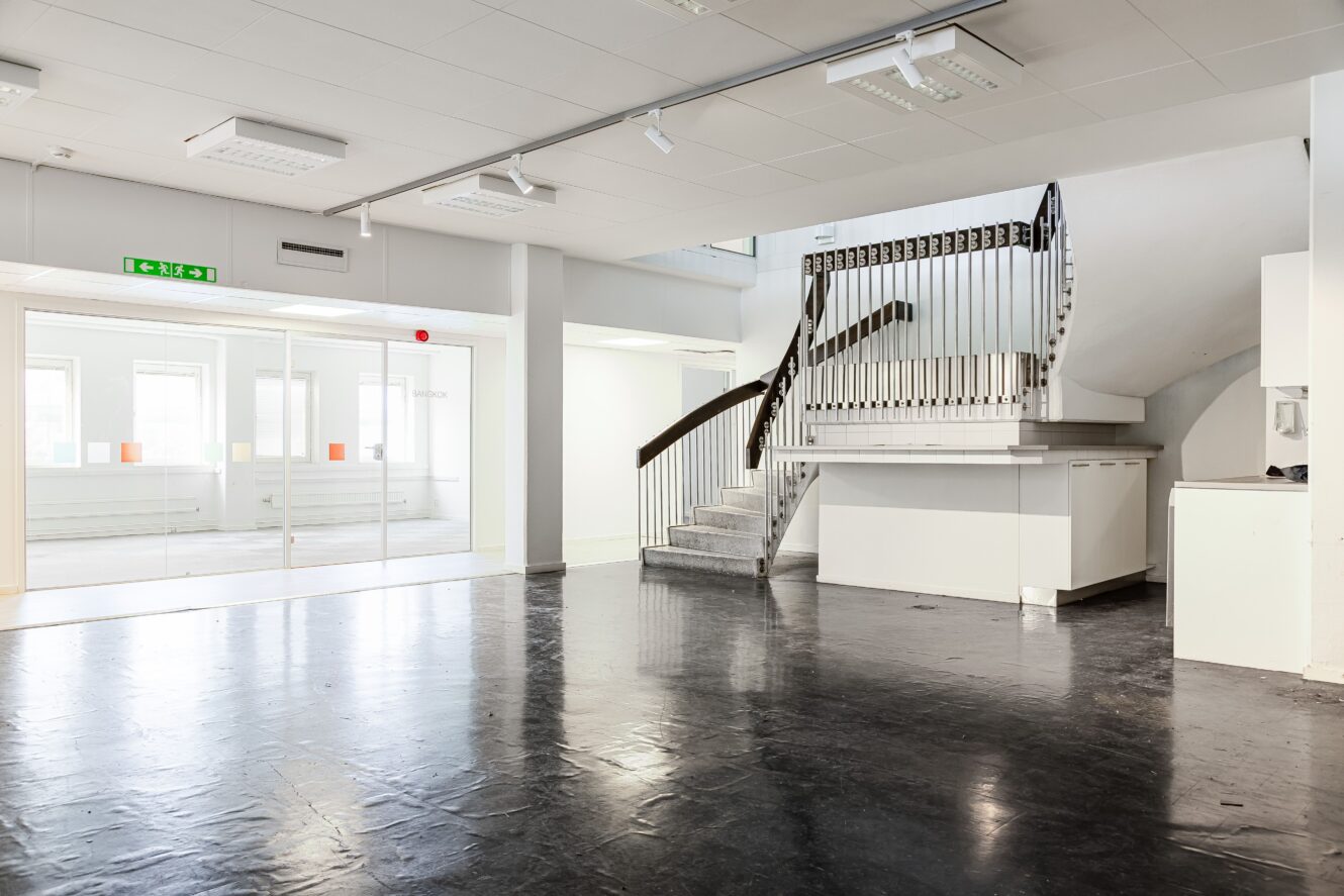 Rymlig, tom interiör med svart golv, vita väggar, en trappa med metallräcke och stora fönster som släpper in naturligt ljus.