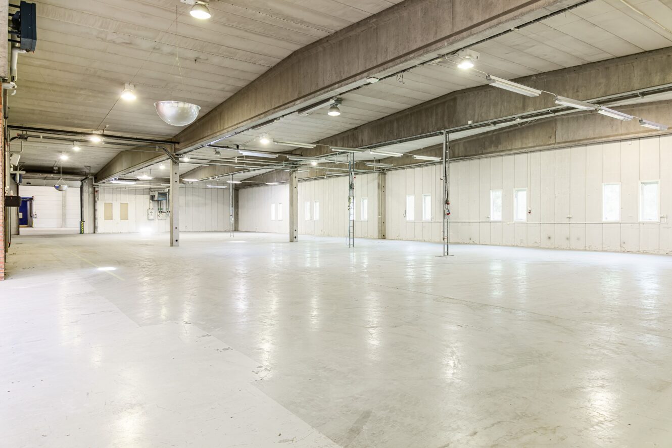 Rymlig, tom lagerlokal med vita golv, högt i tak, stödpelare och flera fönster längs den bortre väggen. Starka taklampor lyser upp interiören.