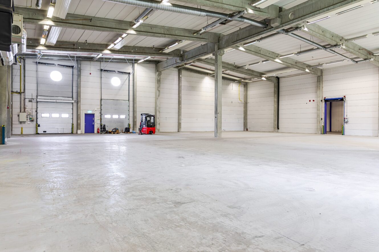 Rymlig, tom lagerlokal med högt i tak, vita väggar, takbelysning, en röd gaffeltruck och lastkajsdörrar längs bakväggen.