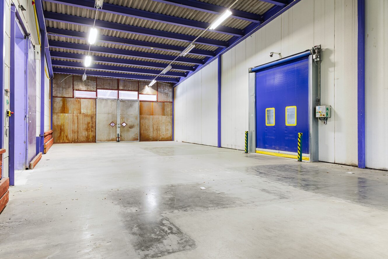 Interiör av ett tomt industrilager med betonggolv, högt i tak, lysrörsbelysning, en stor blå dörr och dubbeldörrar i metall längst bort.