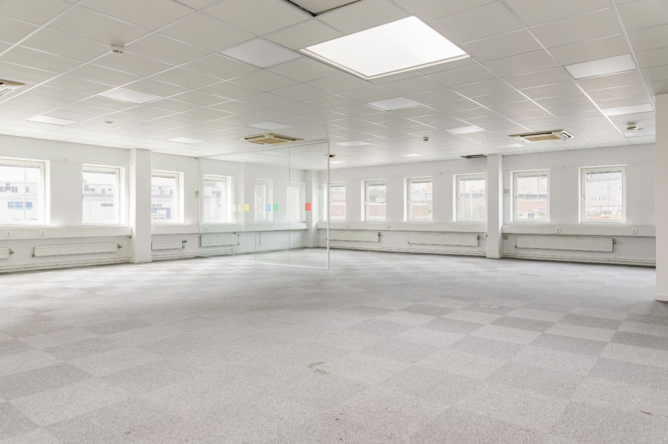 Tomt kontorsutrymme med grå heltäckningsmatta, vita väggar, ett rum med glasväggar i mitten, flera fönster och taklampor.
