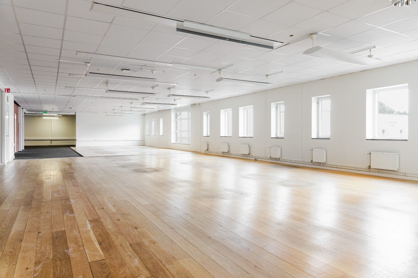 Rymligt, tomt rum med trägolv, vita väggar, taklampor och flera fönster som släpper in naturligt ljus.