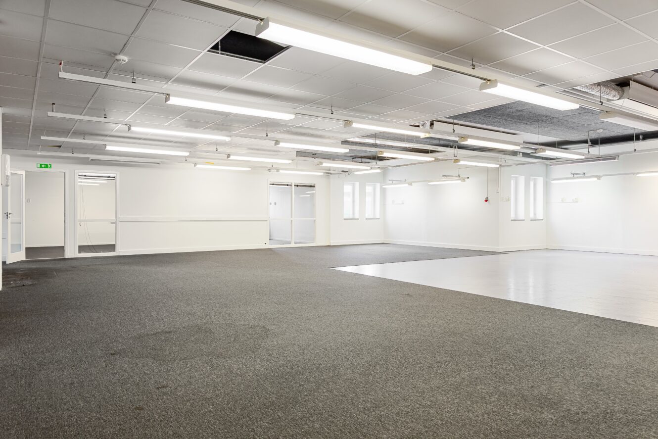 Tomt kontorsutrymme med vita väggar, taklampor, heltäckningsmattor och klinkergolv samt stora fönster som släpper in naturligt ljus.