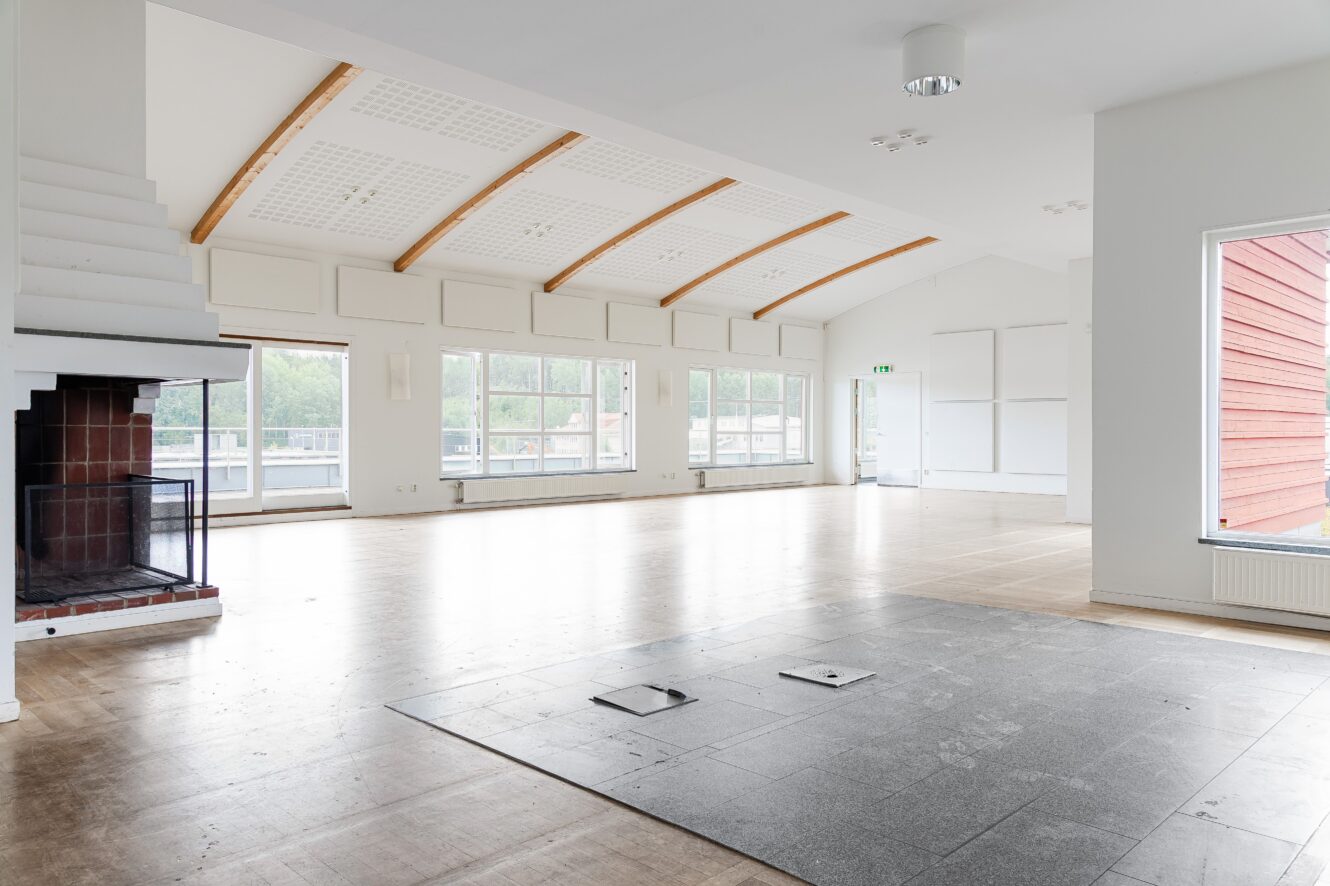 Ett rymligt, tomt rum med stora fönster, en öppen spis till vänster, ljust trägolv och ett grått klinkergolv i mitten. Utrymmet fylls av naturligt ljus.