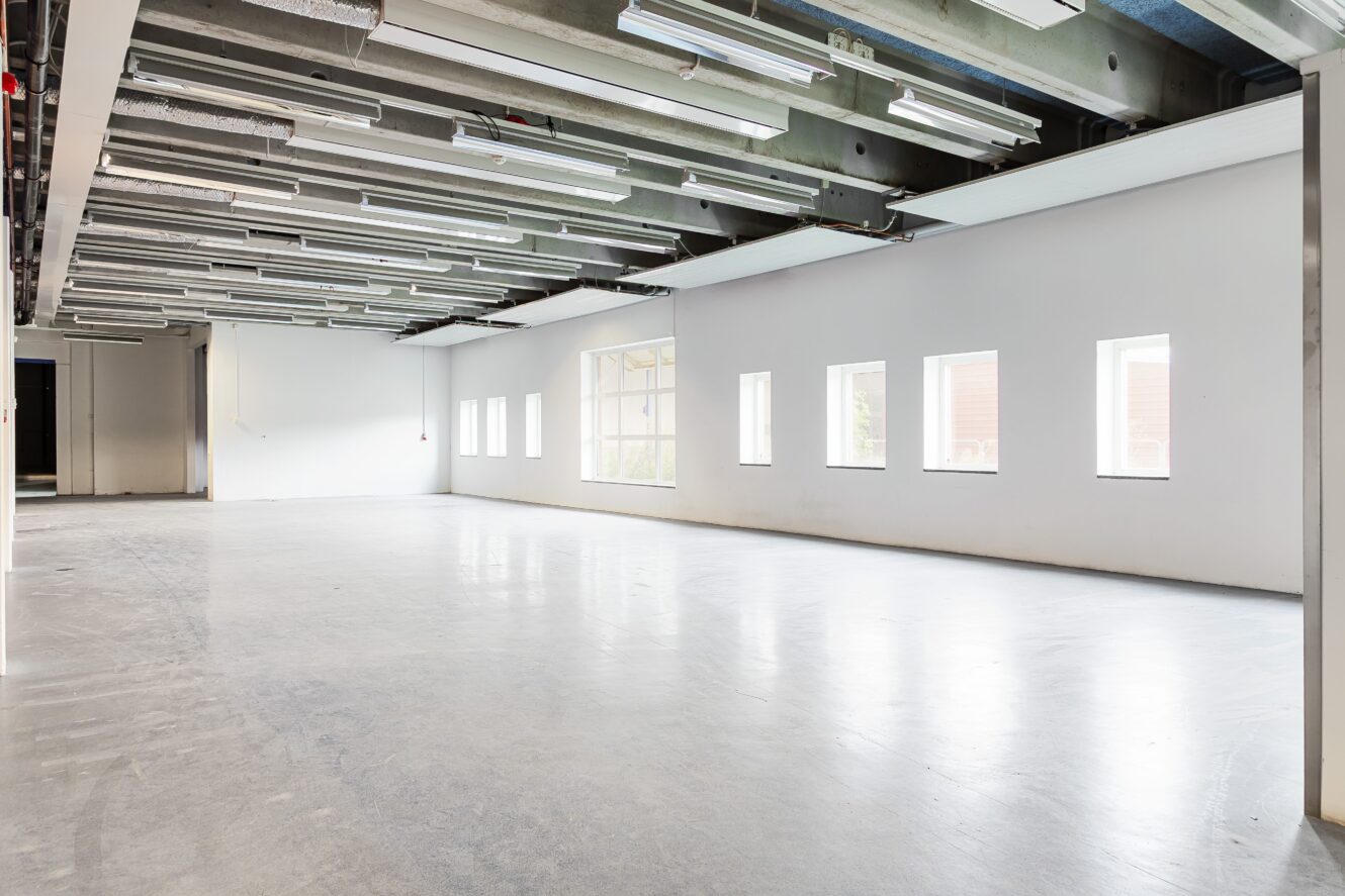 Stort tomt rum med slätt betonggolv, synliga takarmaturer och flera rektangulära fönster längs vita väggar som släpper in naturligt ljus.