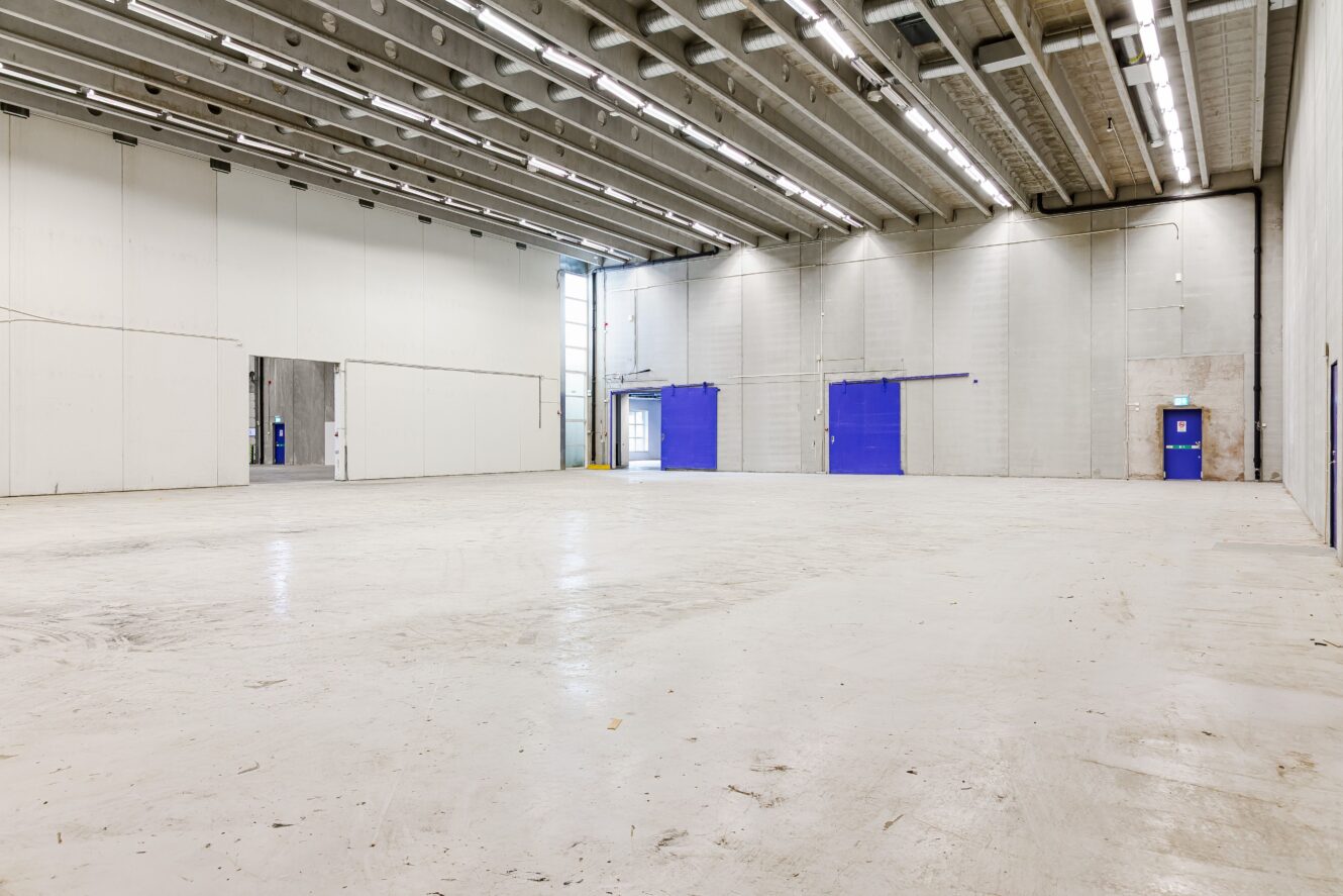 Stort tomt industrilager med vita väggar, högt i tak, synlig belysning och blå dörrar längs ena sidan.