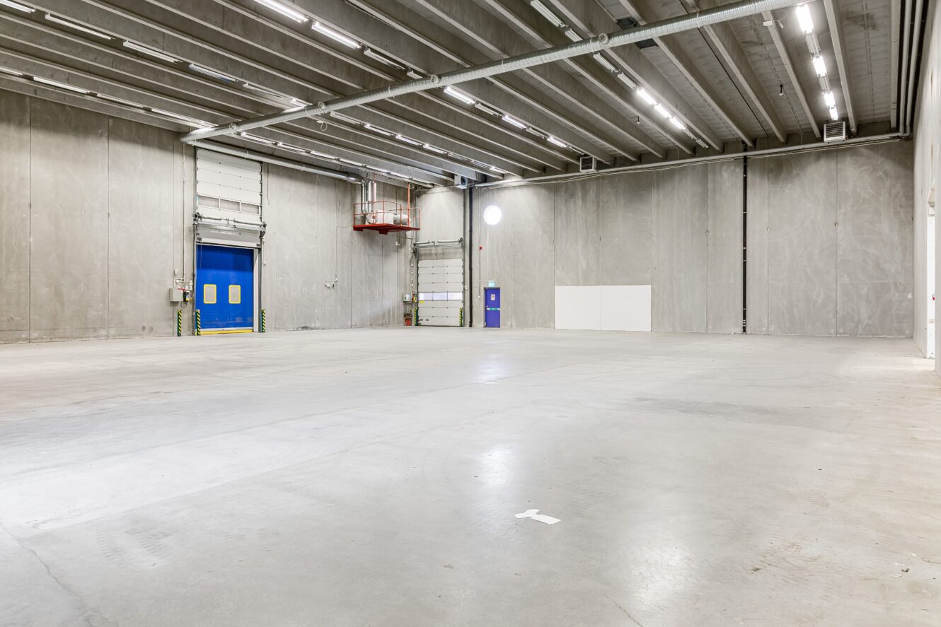 Stort tomt industrilager med betonggolv, högt i tak, synliga bjälkar och en blå lastkajsdörr på den vänstra väggen.