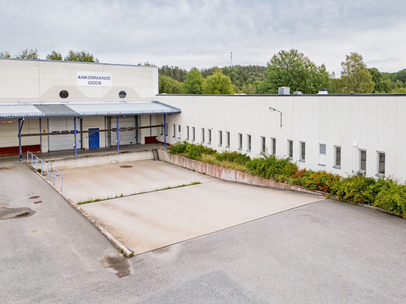 En vit industribyggnad med en lastkaj märkt Ankommande Gods, omgiven av träd och en tom asfalterad yta framför.