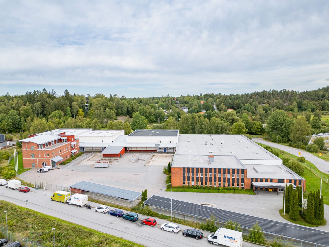 Flygfoto över ett industrikomplex med flera tegelbyggnader, parkeringsplatser, flera parkerade fordon och omgivande gröna träd.