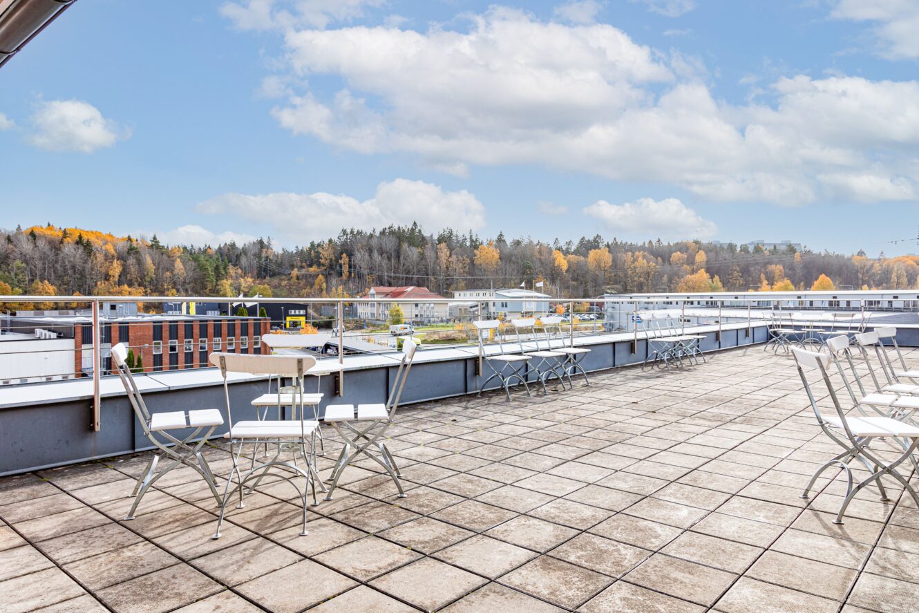 Rymlig takterrass med bord och stolar i metall, utsikt över byggnader och höstträd under en delvis molnig himmel.