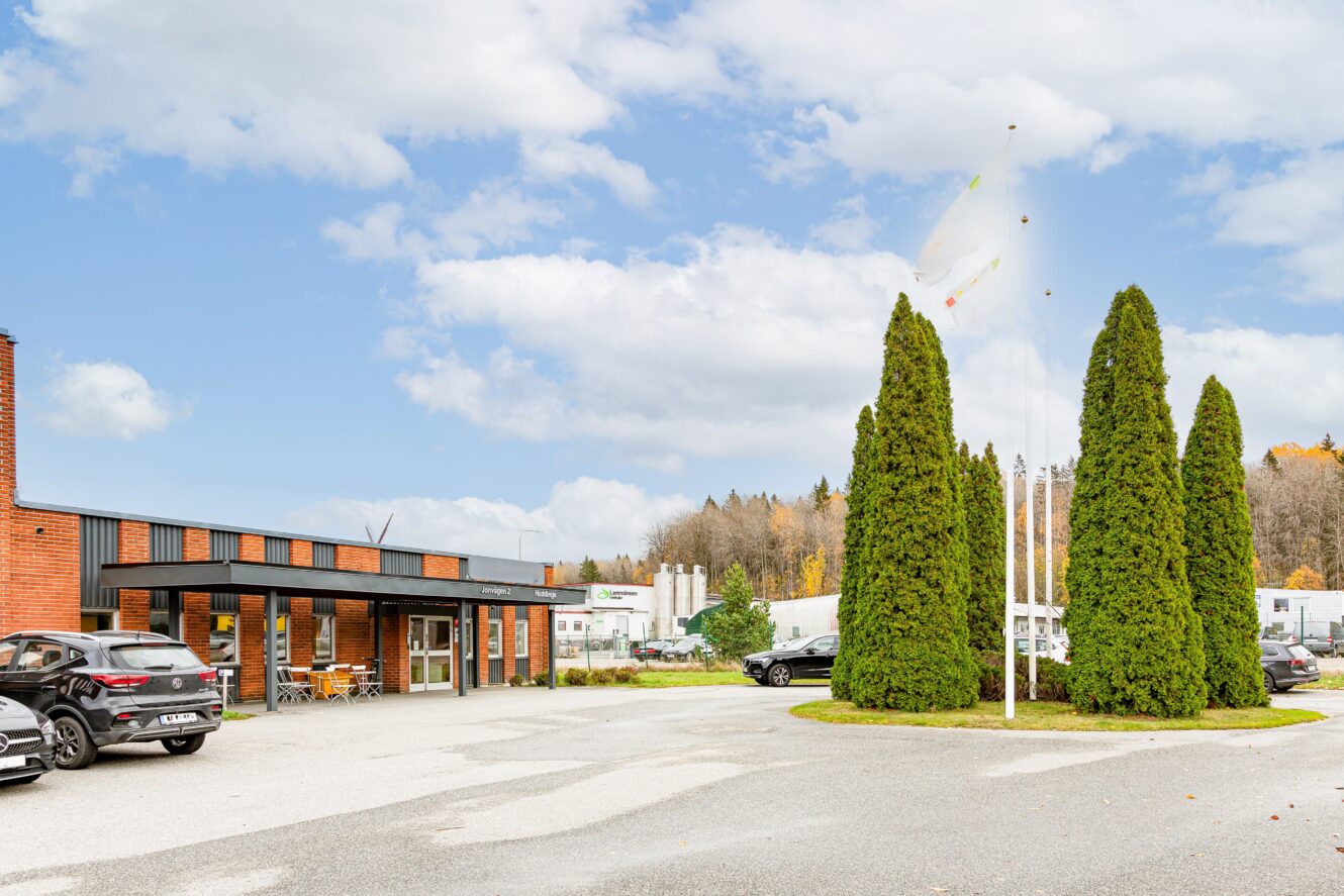 En tegelbyggnad med stora fönster, bilar parkerade utanför, höga gröna träd och vita flaggor på stänger mot en delvis molnig himmel.