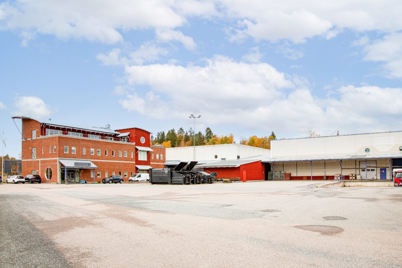 En stor, mestadels tom parkeringsplats framför en röd tegelbyggnad och vita industrihallar under en delvis molnig himmel.