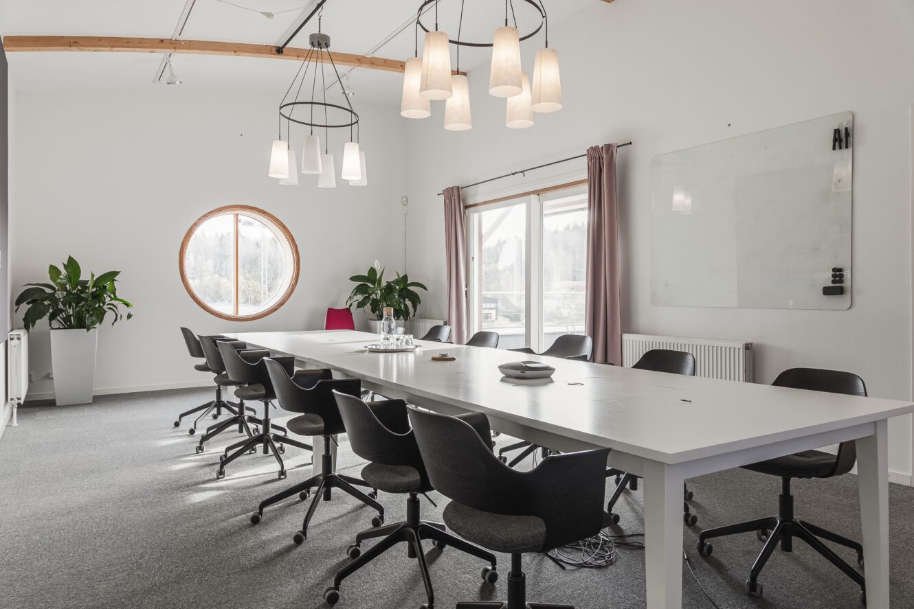 Modernt konferensrum med ett långt vitt bord, svarta stolar, hängande lampor, stora fönster med naturligt ljus, växter och en whiteboard på väggen.
