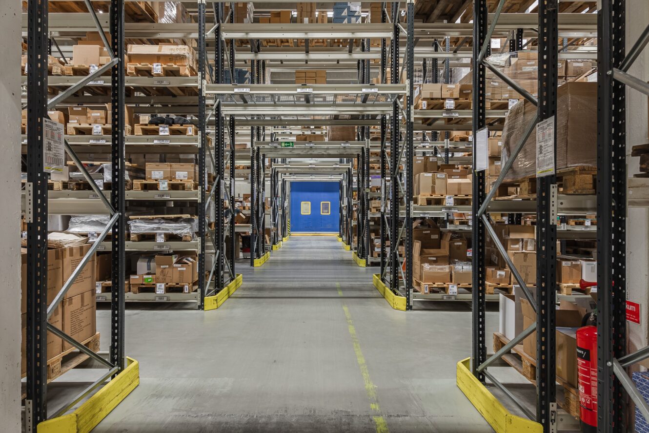 Lagerlokal med höga metallhyllor fyllda med lådor och paket, organiserade längs en bred mittgång som leder till blå dubbeldörrar i bortre änden.