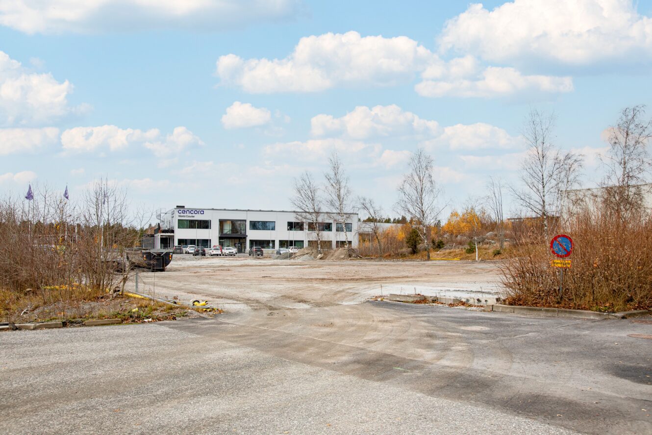 En i stort sett tom grusplan med gles vegetation, några parkerade bilar och en vit industribyggnad i bakgrunden under en delvis molnig himmel.