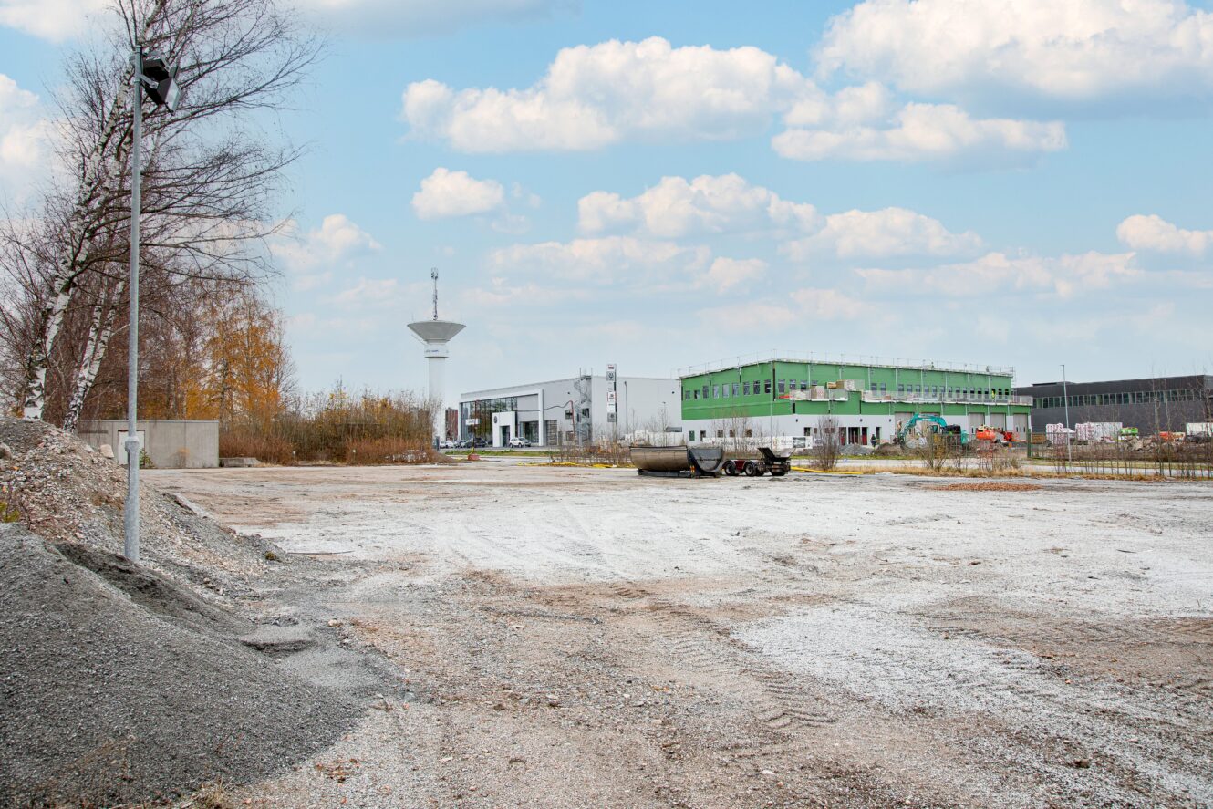 En tom byggarbetsplats med grus och jord i förgrunden, övervakningskameror på stolpar och industribyggnader i bakgrunden under en delvis molnig himmel.