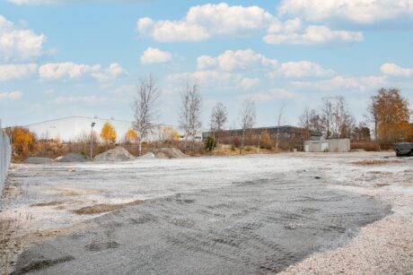 En tom grusplan med glesa träd, grushögar och industribyggnader i bakgrunden under en delvis molnig himmel.