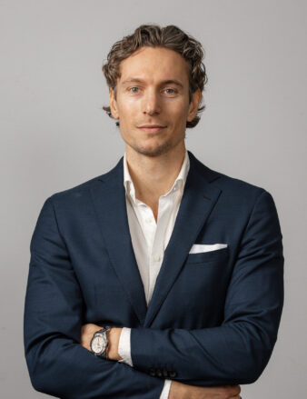 Man i marinblå kostym och vit skjorta står med armarna i kors mot en ljusgrå bakgrund.