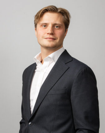 En man i mörk kostym och vit skjorta står framför en ljus bakgrund och tittar in i kameran med ett neutralt ansiktsuttryck.