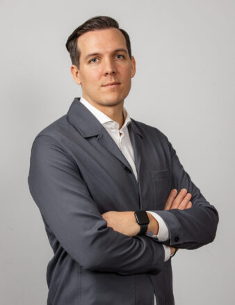 En man med kort mörkt hår, grå kavaj och vit skjorta står med armarna i kors mot en ljus bakgrund.