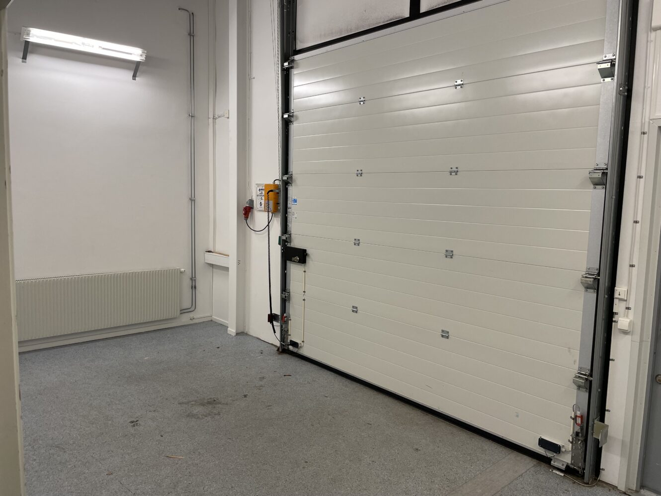 Ett rent, tomt garageutrymme med en stängd vit takskjutport, grått golv, radiator, väggmonterad lampa och elektriska reglage på väggen.