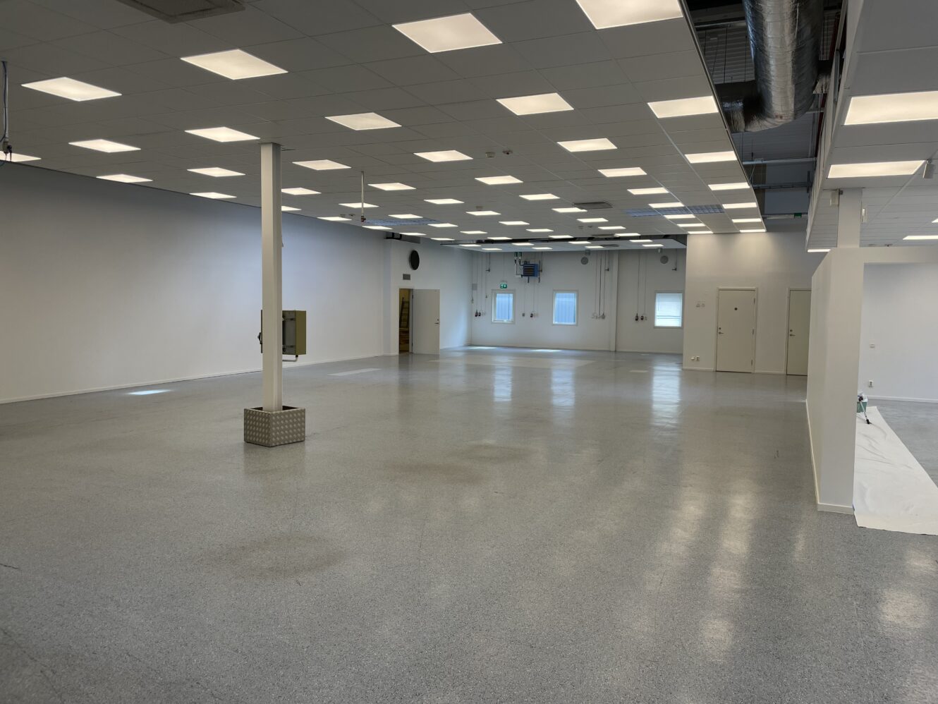 Ett stort, tomt industrirum med vita väggar, gråspräckligt golv, taklampor och synliga kanaler. Flera dörrar och fönster syns längs den bakre väggen.