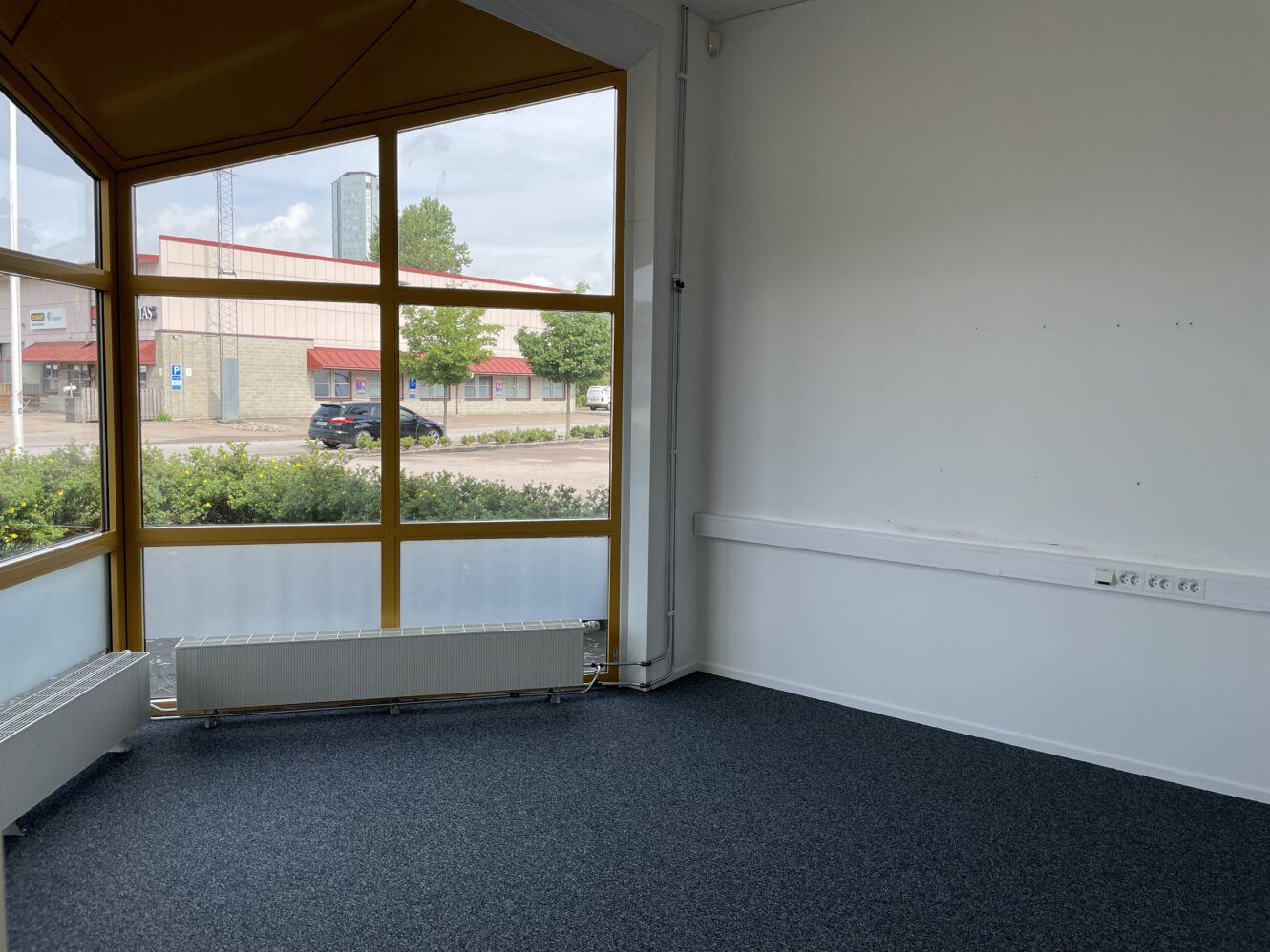 Ett tomt kontorsrum med stora fönster med gul ram, vita väggar, element och grå matta, med utsikt över en gata och kommersiella byggnader utanför.