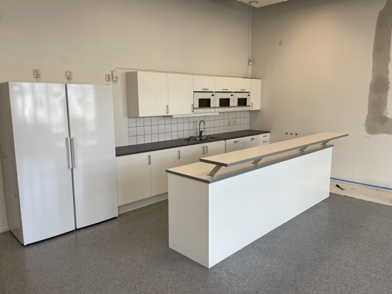 Ett modernt kök med vita skåp, kylskåp med dubbla dörrar, inbyggda mikrovågsugnar, kakelplattor och en vit köksö på ett grått golv.