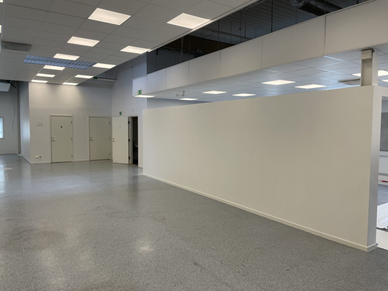 Rymligt, tomt rum med vita väggar, grått golv, flera stängda dörrar och taklampor; delvis andra våningen med räcke ovanför.