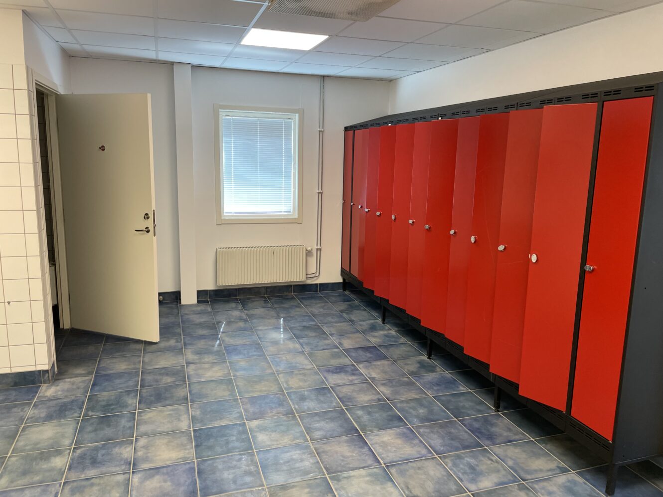 Ett omklädningsrum med röda skåp, blått klinkergolv, vita klinkerväggar, ett fönster med persienner och en öppen dörr.