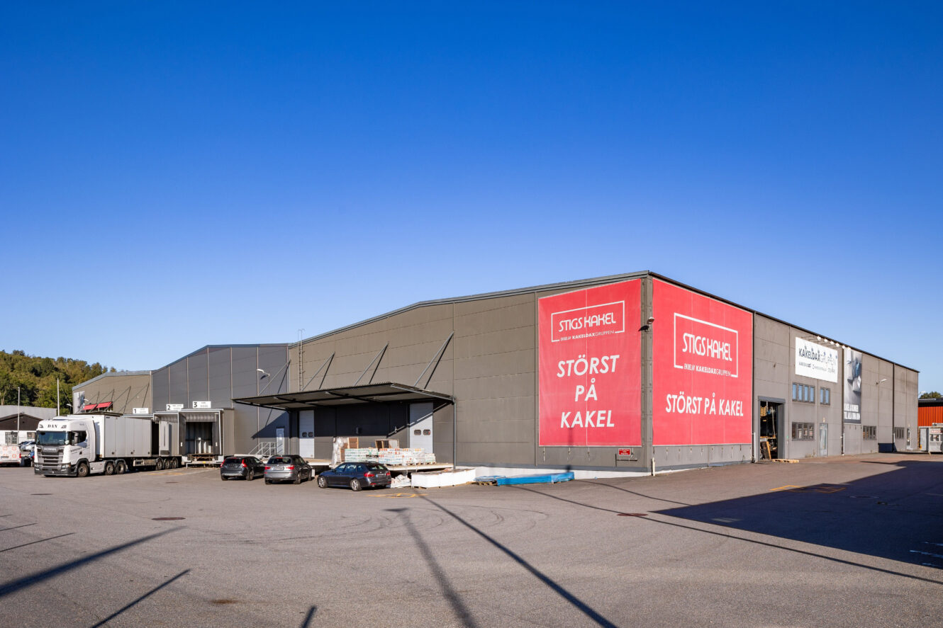 Stor kommersiell lagerbyggnad med grå fasad, röd skylt med texten STÖRST PÅ KAKEL, parkerade bilar, en lastbil och klarblå himmel.