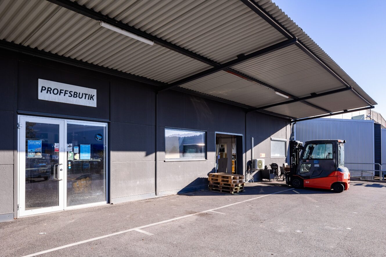 En gaffeltruck står parkerad vid lastpallar utanför en grå byggnad märkt PROFFSBUTIK med glasdörrar och fönster under en markis av korrugerad plåt.