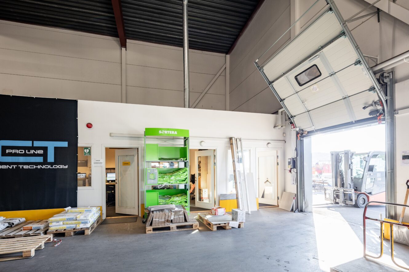 Interiör i lagerlokal med industriutrustning, öppen garageport, staplade pallar och en gaffeltruck som syns utanför i solljus.