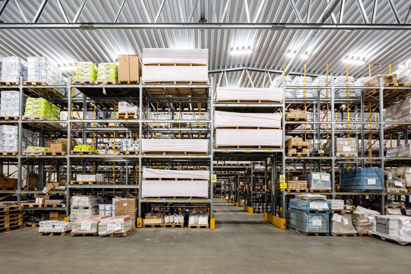 Stor lagerlokal med höga metallhyllor staplade med olika lådor, pallar och förpackade varor under stark industriell belysning.