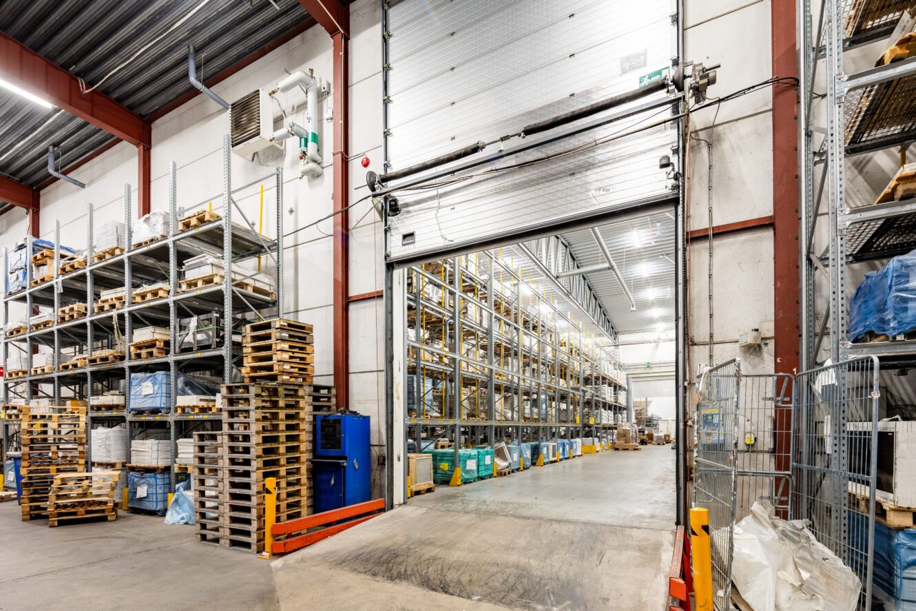 Industriell lagerlokal med höga metallhyllor där pallar och olika varor förvaras, en stor öppen lastbryggedörr och betonggolv.