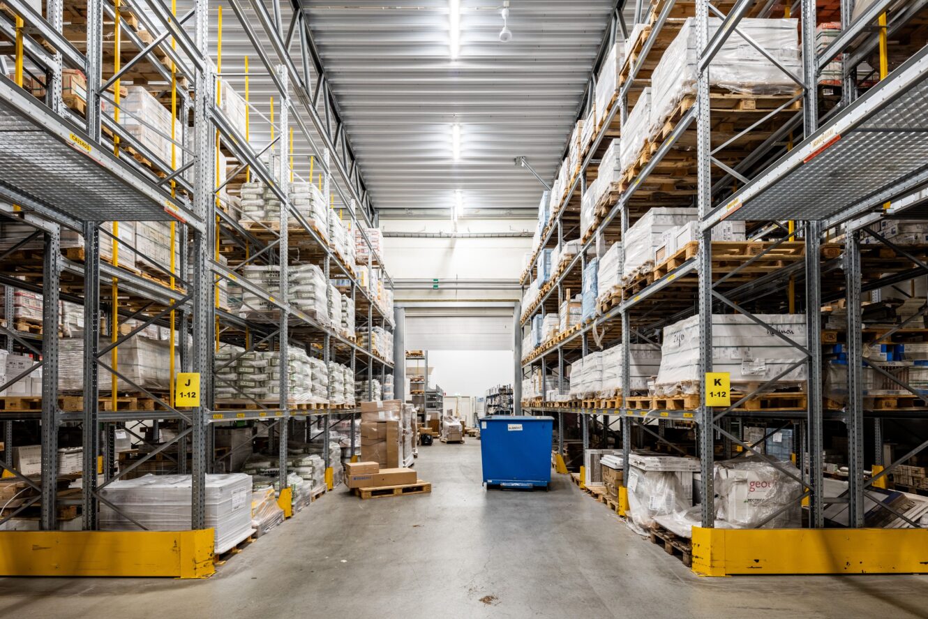 Stor lagerlokal med höga metallhyllor fyllda med pallar och lådor, stark takbelysning och en blå vagn i mittgången.